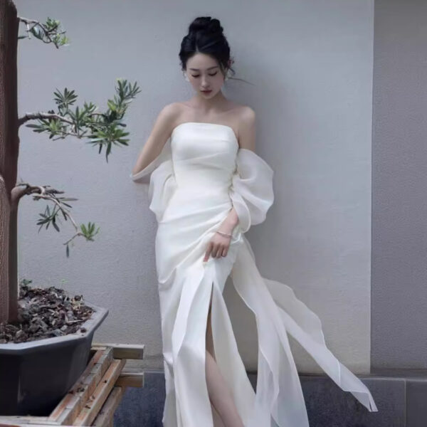 Elegant woman in a strapless white gown with flowing sleeves, posing beside a potted plant against a minimalist background. The dress features a thigh-high slit, showcasing a sophisticated bridal or formal look.