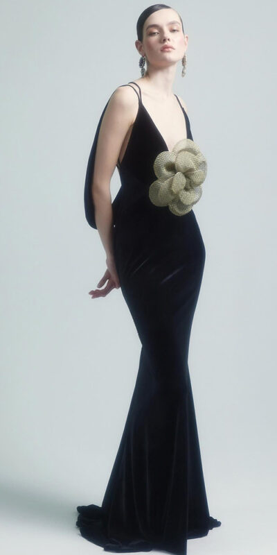 Elegant model wearing a black velvet evening gown with a unique oversized flower embellishment at the waist, set against a soft gray background. The dress features thin straps and a mermaid silhouette, showcasing a sophisticated and modern design.