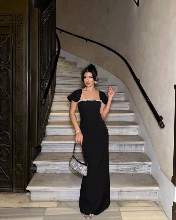 Elegant woman in a stylish black evening gown stands gracefully on a marble staircase, holding a sparkling handbag. The setting features a sophisticated architectural backdrop, enhancing the glamorous ambiance of the scene.