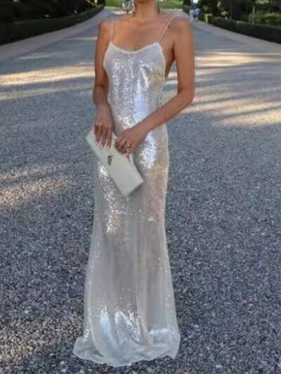 Elegant woman wearing a shimmering silver sequin gown with thin straps, holding a stylish clutch, standing on a gravel pathway surrounded by greenery.