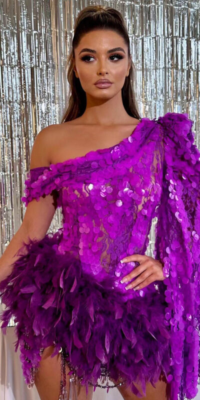 Model in a vibrant purple dress adorned with sequins and feathers, posing against a shimmering silver backdrop, showcasing a stylish one-shoulder design.