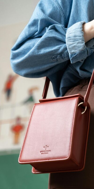 Close-up of a stylish red leather sling bag worn over a rolled-up denim shirt, indoors.