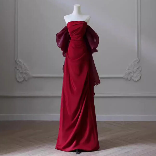 Elegant red evening gown displayed on a mannequin, featuring a strapless design and flowing sheer sleeves. The dress is styled in a sophisticated manner, set against a minimalist background with light gray walls and wooden flooring, highlighting its luxurious fabric and silhouette. Ideal for formal events and special occasions.