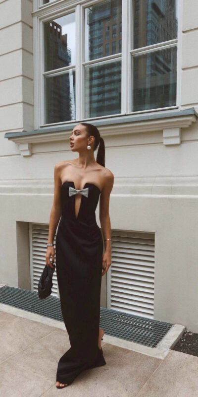 A stylish woman wearing a black strapless evening gown with a bow detail, standing against a modern building backdrop. She accessorizes with statement earrings and a black clutch, showcasing a chic urban fashion look.