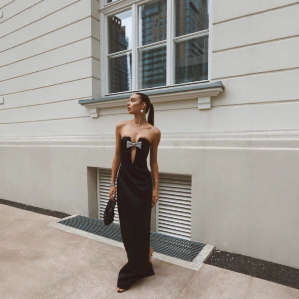 A stylish woman wearing a black strapless evening gown with a bow detail, standing against a modern building backdrop. She accessorizes with statement earrings and a black clutch, showcasing a chic urban fashion look.