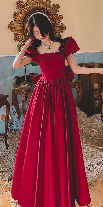 Elegant woman in a red ball gown with puff sleeves, styled with a necklace, standing in a vintage room with antique furniture and decorative accents.