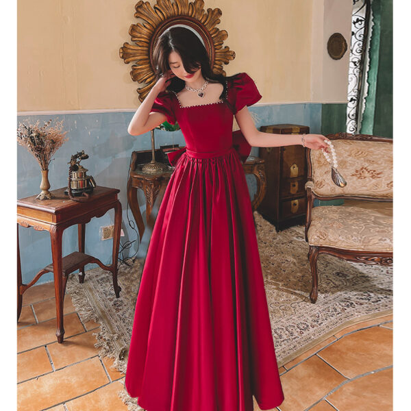 Elegant woman in a red ball gown with puff sleeves, styled with a necklace, standing in a vintage room with antique furniture and decorative accents.