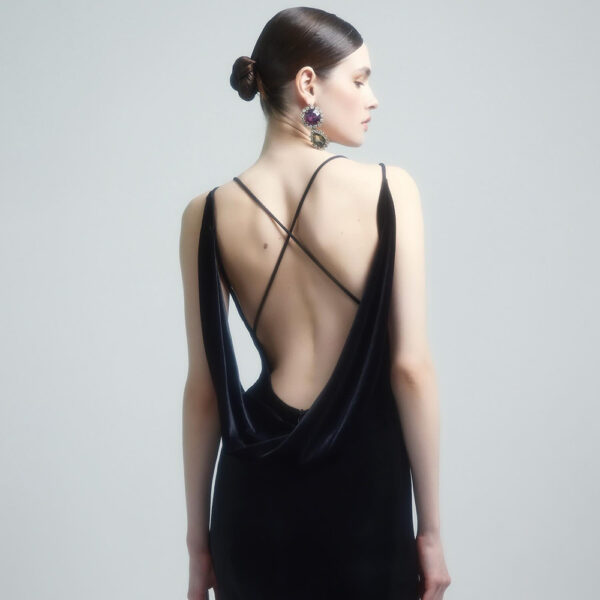 Model showcasing a stylish black velvet dress with a deep back and crisscross straps, complemented by large, colorful earrings, set against a soft gray background.