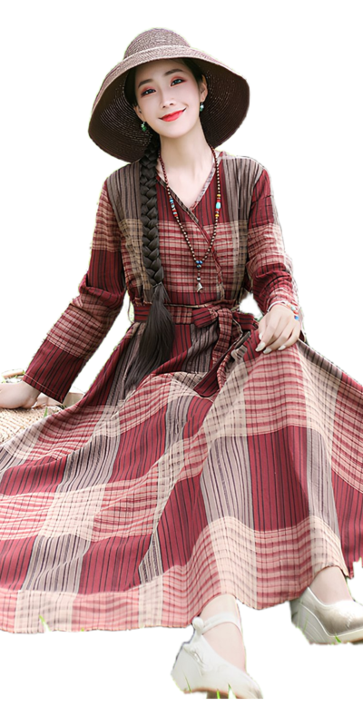 Woman in a stylish plaid maxi dress with a belt, seated outdoors, wearing a wide-brimmed hat and accessorized with a necklace. The dress features a mix of warm colors and flowing fabric, perfect for a casual summer look.