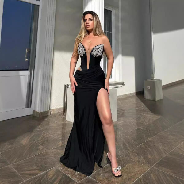 Elegant woman in a black evening gown with a plunging neckline and floral embellishments, posing confidently with one leg exposed. The setting features modern architecture and natural lighting, highlighting the dress's sophisticated design.