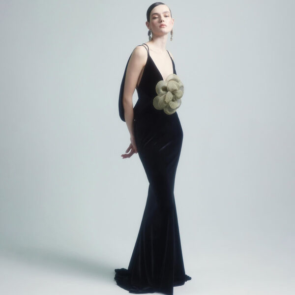 Elegant model wearing a black velvet evening gown with a unique oversized flower embellishment at the waist, set against a soft gray background. The dress features thin straps and a mermaid silhouette, showcasing a sophisticated and modern design.