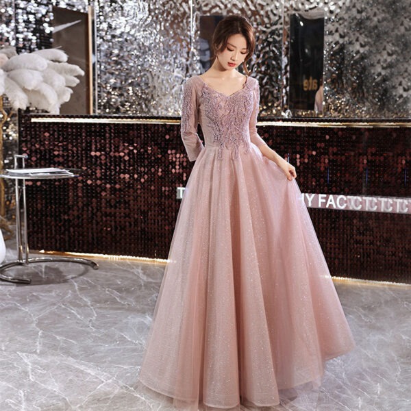 Elegant woman in a blush pink evening gown with intricate beadwork, standing gracefully in a stylish, modern setting with a shimmering backdrop. The gown features long sleeves and a flowing skirt, perfect for formal events or special occasions.