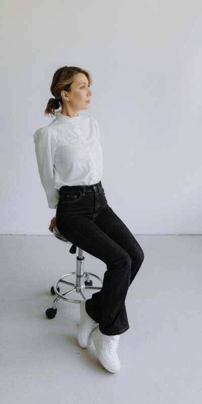 Elegant woman in white blouse and black pants posing on stool in studio setting, showcasing modern fashion.