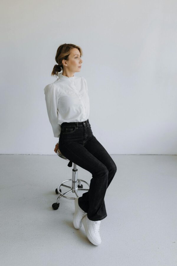 Elegant woman in white blouse and black pants posing on stool in studio setting, showcasing modern fashion.