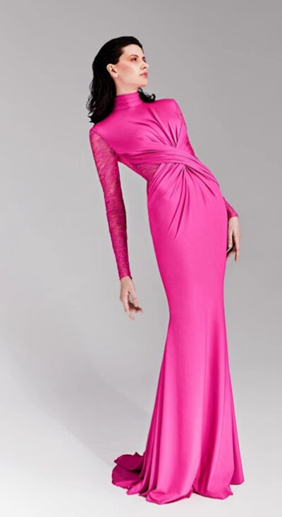 Model wearing a vibrant pink evening gown with a high neckline and one long lace sleeve, showcasing elegant draping and a fitted silhouette against a neutral background.