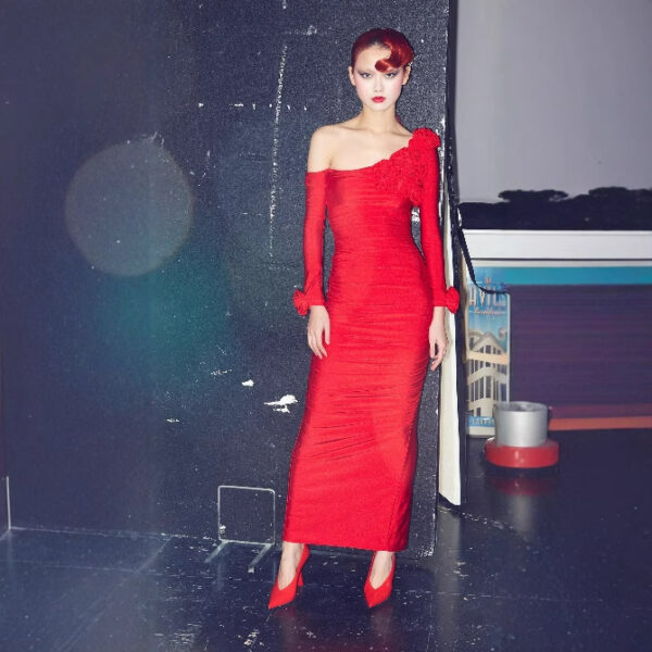 Fashion model wearing a striking red off-shoulder dress with a floral detail, posing elegantly against a dark background. The outfit is complemented by matching red high heels, showcasing a bold and sophisticated style.