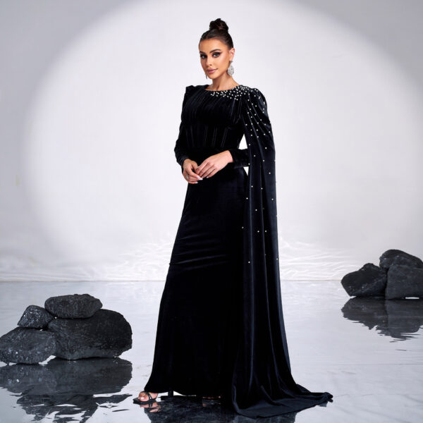 Elegant woman in a black velvet gown with a structured design and embellished shoulder cape, posing against a minimalist backdrop with reflective surfaces and dark stones. The outfit features intricate detailing and a sophisticated silhouette, perfect for formal occasions.