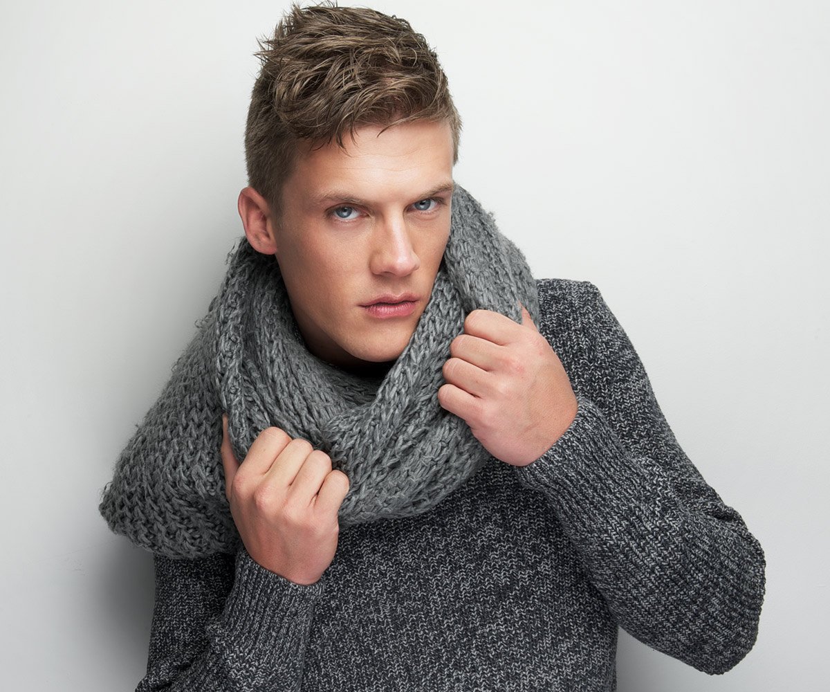 Young man wearing a chunky gray knit scarf and a textured gray sweater, posing against a neutral background. His expression is confident and engaging, showcasing winter fashion styles.